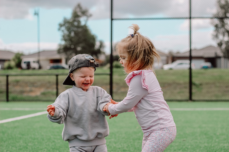 toddler boy in grey and toddler girl in pink holding hands on a sports field