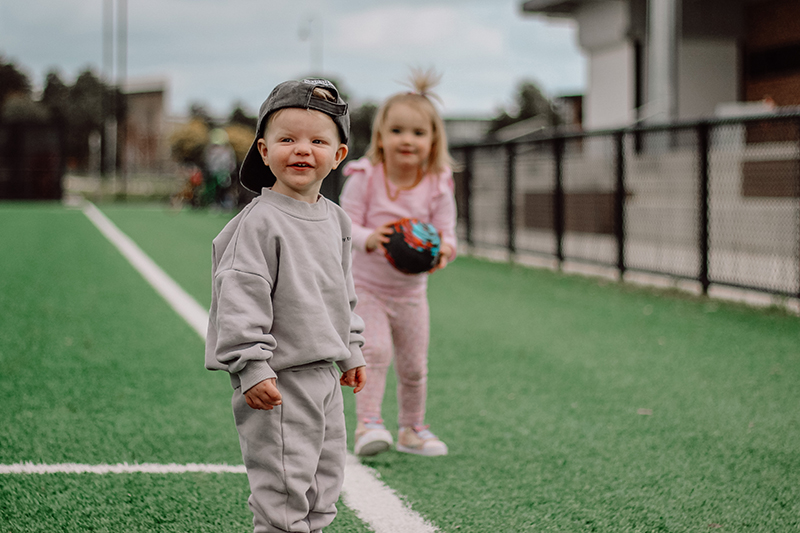 toddler boy in grey on sports field with toddler girl in pink in background