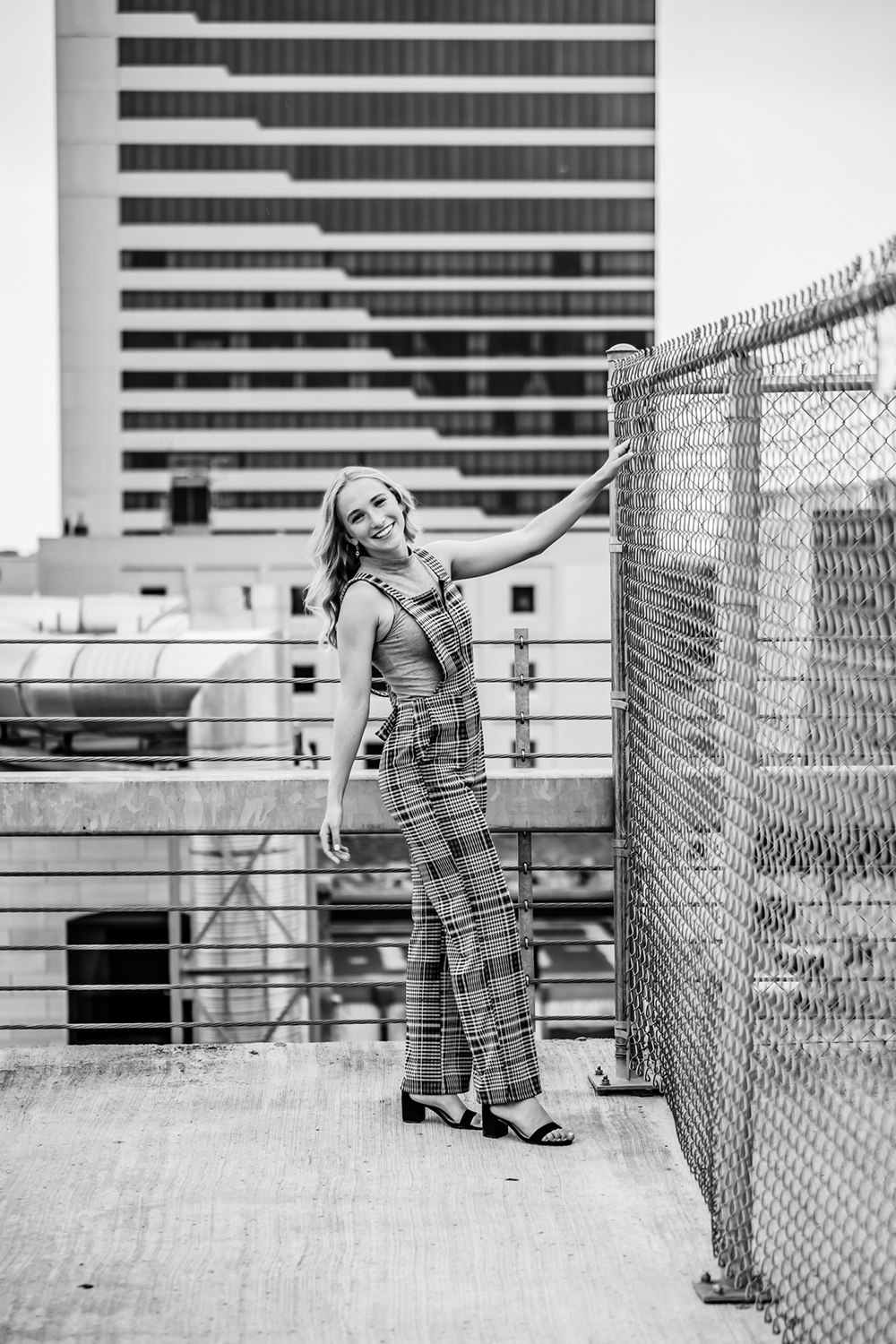 teenage girl posing on a rooftop holding onto a wire fence with city skyline in the background
