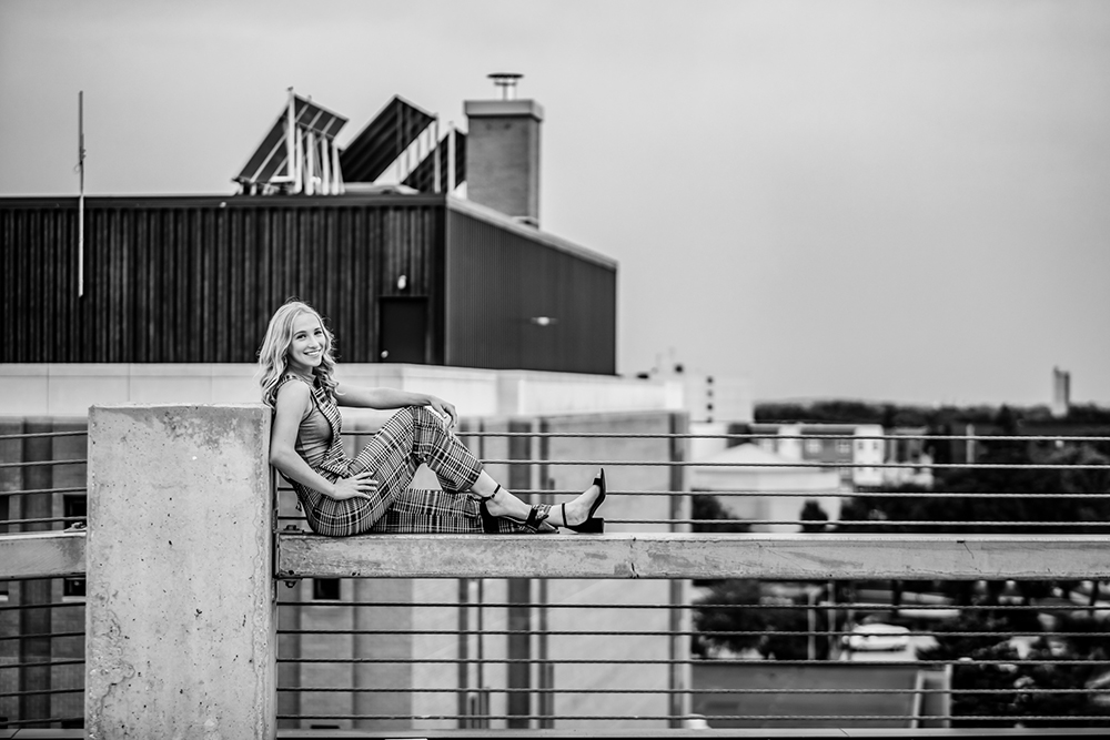 Black and white photo of a senior girl posing on a fence with city skyline in the background