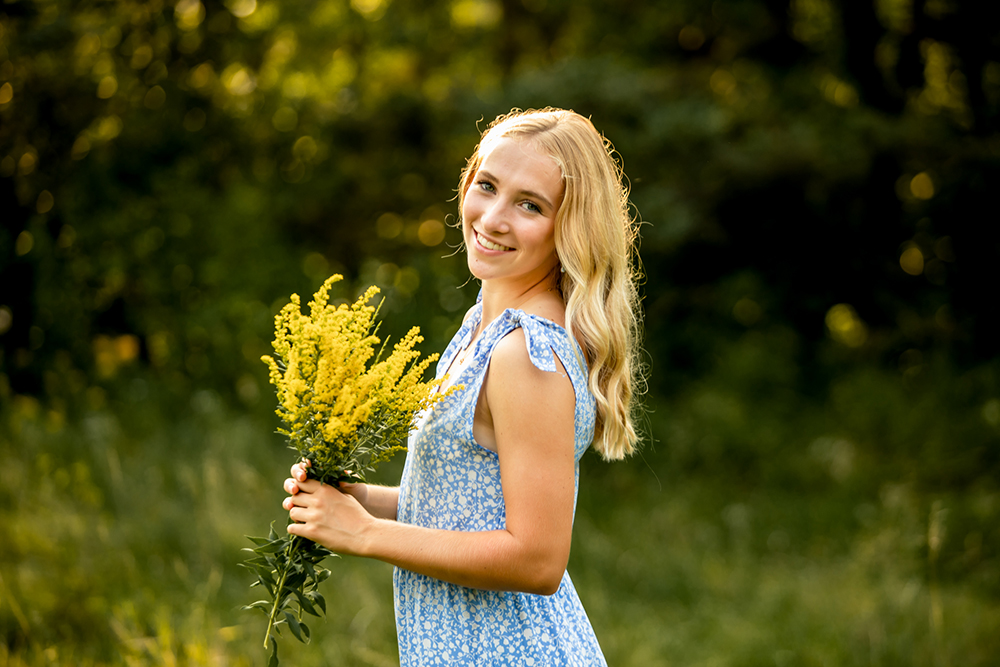 teen girl poses with long blonde hair holding yellow flowers and wearing a blue dress