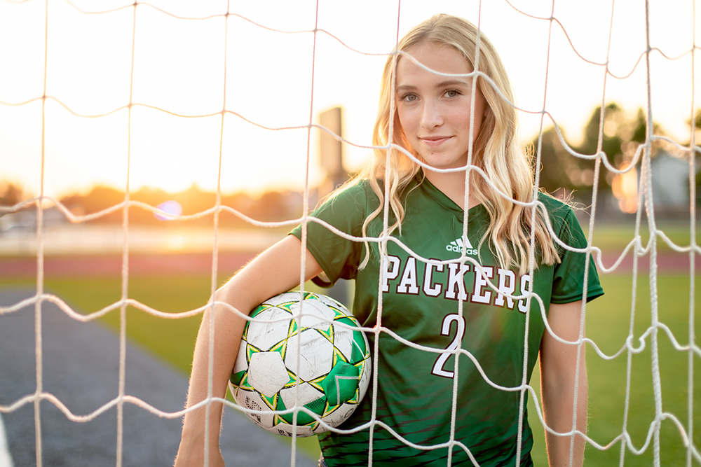 senior girl holding a soccer ball and standing behind a soccer net