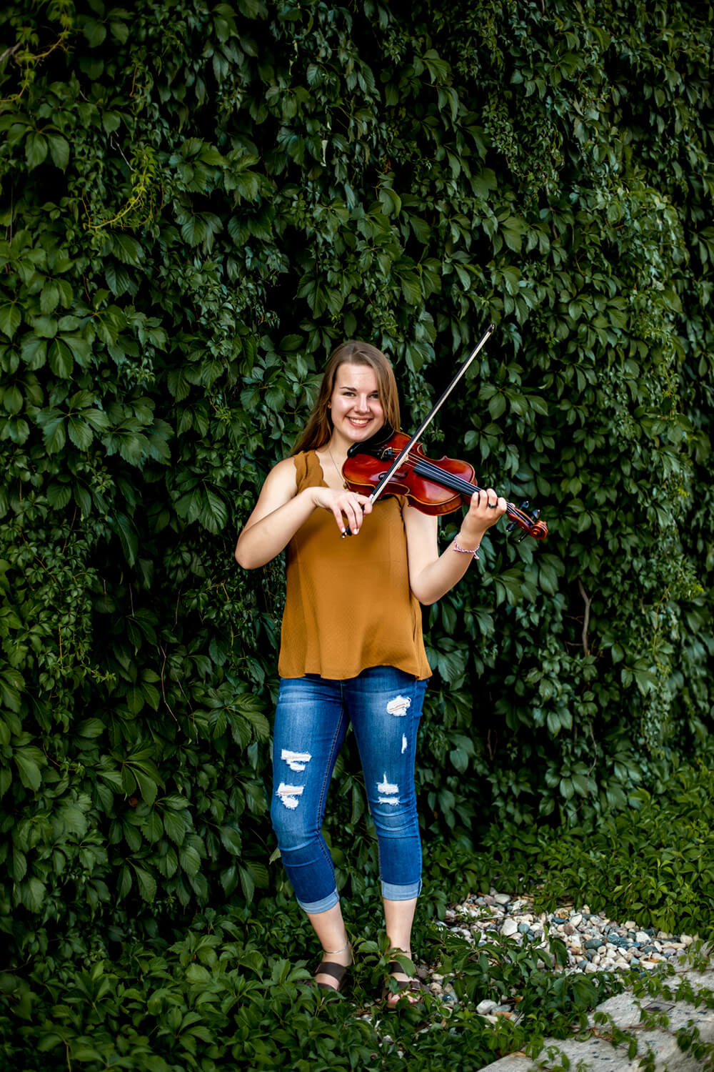girl standing in front of an ivy covered wall playing a violin.
