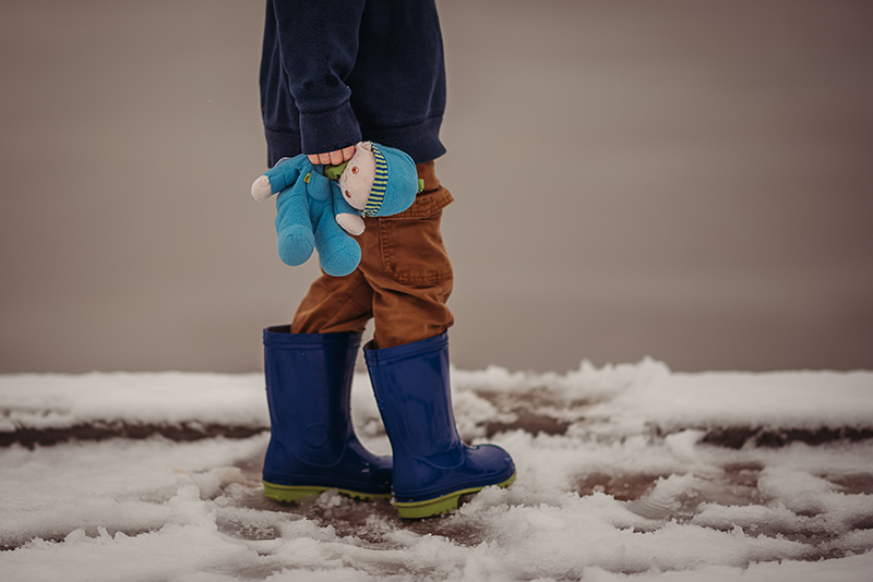 small child walking in the snow wearing blue boots and holding a soft toy
