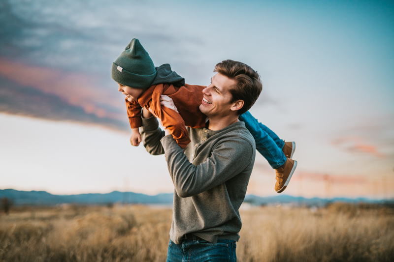 father holding toddler up against sunset sky